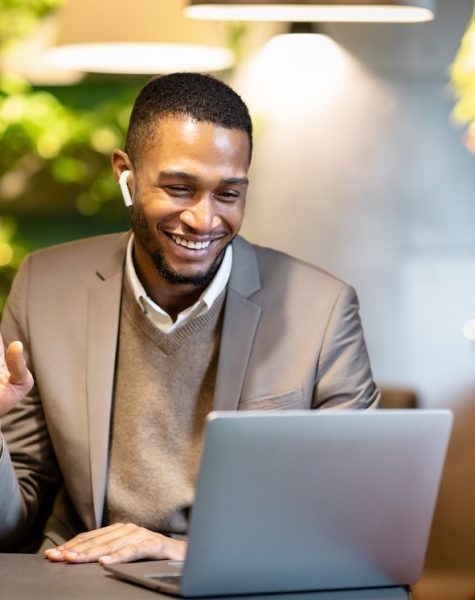 Smiling black business man making video call to family Coffee Break. Happy afro businessman in earbuds making video call to family using laptop at cafe, waving hand, copyspace